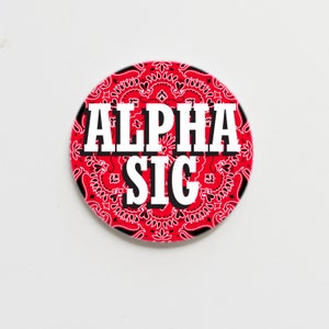 May include: A red and black circular button with a white bandana pattern and the text "ALPHA SIG" in white.