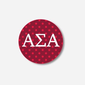 May include: A circular button with a maroon base and small red polka dots. The button displays the Greek letters ΑΣΑ in white. The button has a shadow, creating a 3D appearance. The design is simple and bold.