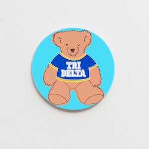 May include: A brown teddy bear wearing a blue shirt with the text "TRI DELTA" in white and yellow letters on a light blue background.