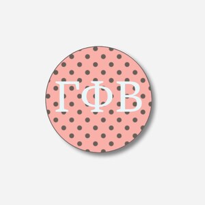 May include: A round, pink button with a polka dot pattern and the Greek letters ΓΦB in white. The button has a dark brown border and a shadow effect, giving it a three-dimensional appearance.
