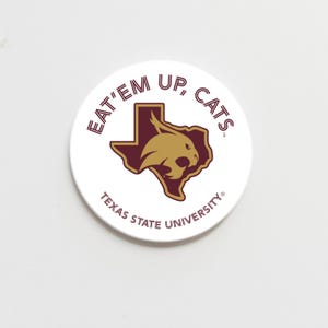 May include: A white circular button with the text "EAT'EM UP, CATS" in maroon. The button features a maroon outline of the state of Texas with a gold bobcat head in the centre. The text "TEXAS STATE UNIVERSITY" is also present.