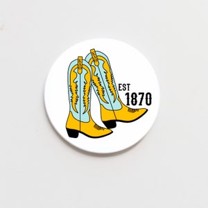 May include: White round sticker with a blue and yellow illustration of two cowboy boots. The text "EST 1870" is printed in black.