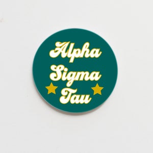 May include: A teal blue round sticker with white text that reads "Alpha Sigma Tau" in a retro font. Two gold stars are on either side of the text.