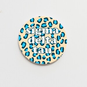 May include: A round coaster with a light beige background and a blue and black leopard print pattern. The text "sigma delta tau" is written in white on the coaster.