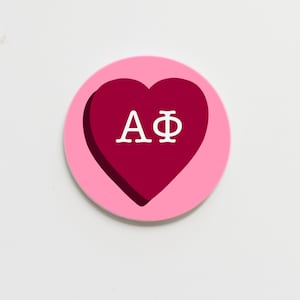 May include: A circular, pink badge featuring a dark red heart shape at its centre. The heart displays the white Greek letters A and Φ. The badge has a shadow effect, creating a 3D look.