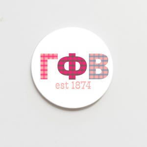 May include: A white round coaster with a pink, red, and blue plaid design that spells out "ΓΦΒ est 1874".