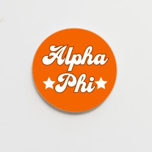 May include: Orange round sticker with white text that reads "Alpha Phi" in a retro font with two white stars on either side of the text.