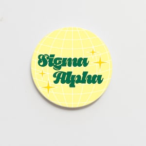 May include: A round, yellow coaster with a grid pattern and the words "Sigma Alpha" in green script. Four yellow starbursts are scattered around the text, creating a retro design.