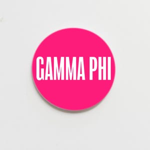 May include: A round, hot pink button with the white text "GAMMA PHI" centered on the face. The button is set against a plain white background.