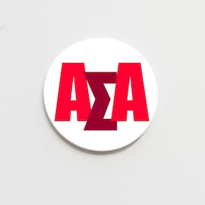 May include: A white, circular coaster with the Greek letters ΑΣΑ in red and maroon. The letters are prominently displayed in a bold font. The coaster's design is simple and eye-catching, suggesting it's made from ceramic or a similar material.