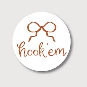 May include: A round white sticker with a brown bow illustration at the top and the words "hook 'em" in brown script below. The sticker has a simple, clean design.