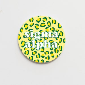 May include: A round sticker with a yellow and green leopard print design. The text "sigma alpha" is written in white letters on the sticker.