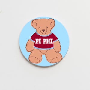 May include: A light blue round coaster with a cartoon illustration of a brown teddy bear wearing a red shirt with the text "PI PHI" in white.