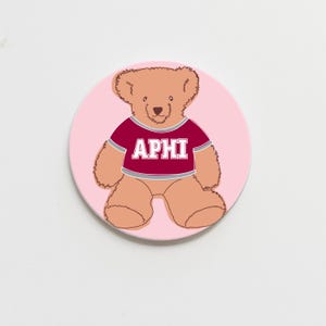 May include: A pink and white round coaster with a brown teddy bear wearing a maroon shirt with the letters "APHI" printed in white.