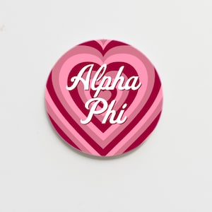 May include: A pink and red heart-shaped button with the text "Alpha Phi" in white cursive lettering.