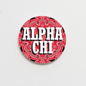 May include: A red and black circular button with a white bandana pattern and the text "ALPHA CHI" in white.