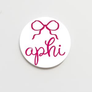 May include: White round coaster with a pink bow and the word "aphi" written in cursive.