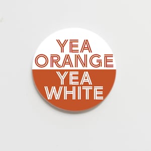 May include: A round button with a white top half and an orange bottom half. The text "YEA ORANGE YEA WHITE" is printed in white, stylized font across the button.