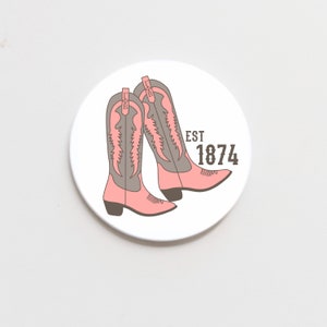 May include: A white round coaster with a pink and brown illustration of two cowboy boots. The text "EST 1874" is printed on the coaster.