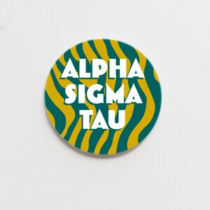 May include: A round, yellow and green button with a white zebra stripe pattern. The text "ALPHA SIGMA TAU" is printed in white on the button.