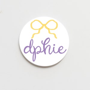 May include: A white round coaster with a yellow bow and the name "dphie" written in purple.