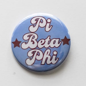 May include: A blue button with white text that reads "Pi Beta Phi" in a retro font. Two brown stars are on either side of the text.