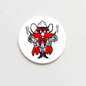 May include: A white, circular coaster featuring a cartoon cowboy with a red mustache, beard, and cape, holding two black pistols. The cowboy wears a black hat and vest. The design is in black and red.