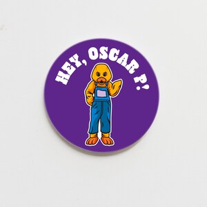 May include: A round, purple button with the text "HEY, OSCAR P!" in white. The button features a cartoon duck wearing blue overalls, orange feet, and a yellow body. The duck is waving.