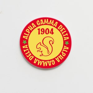 May include: A red and yellow round sticker with the text "Alpha Gamma Delta" and "1904" in a yellow circle with a green star on each side. The circle also features a yellow squirrel silhouette.
