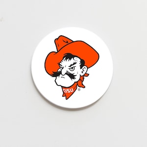 May include: White round coaster featuring the Oklahoma State University (OSU) mascot, a cowboy with an orange hat and bandana. The cowboy has a black mustache and the OSU logo is on the bandana.