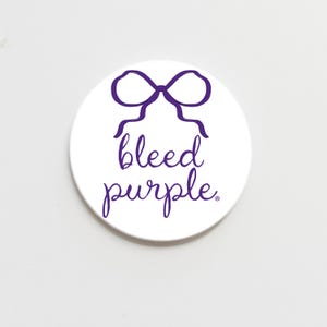 May include: A white round button with the phrase "bleed purple" in a purple script font. Above the text is a purple bow illustration. The button is on a white background.
