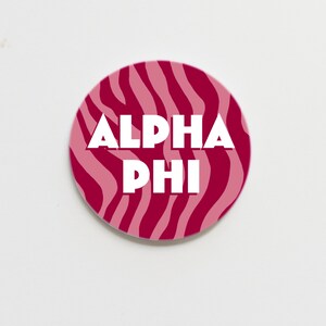 May include: A pink and white zebra print round sticker with the text "ALPHA PHI" in white.