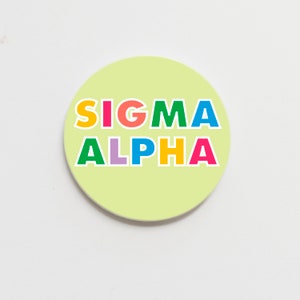 May include: A round, light yellow coaster with the text "SIGMA ALPHA" in colourful letters. The letters are in different colours: S is yellow, I is pink, G is green, M is blue, A is red, A is yellow, L is light blue, P is yellow, H is pink, and A is red.