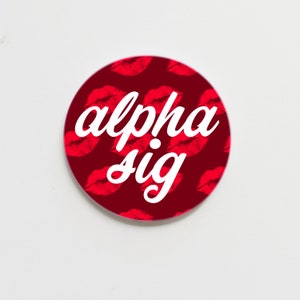 May include: A round button with a red background and a white pattern of lips. The text "alpha sig" is written in white cursive font.