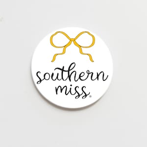May include: A white circular item featuring a yellow bow design at the top and the words "southern miss" in black cursive font. The design is centered on the white background.