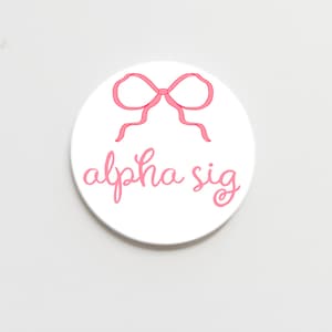 May include: White round coaster with a pink bow and the text "alpha sig" in pink cursive lettering.