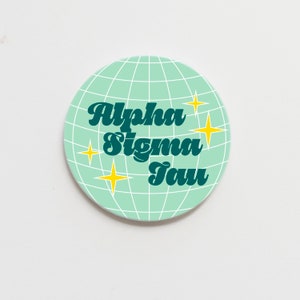 May include: A circular, pale green coaster featuring a white grid design, like a globe. Teal script spells out "Alpha Sigma Tau". Four yellow starbursts are placed around the text.