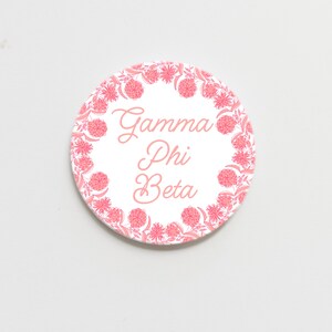 May include: A round, white button with the words "Gamma Phi Beta" in pink script. The button is encircled by a wreath of pink flowers and leaves, creating a decorative border. The design is simple and elegant.