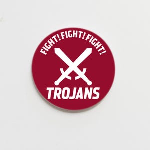 May include: A red round button with white text that says "FIGHT! FIGHT! FIGHT!" and "TROJANS" with a white crossed swords graphic in the center.