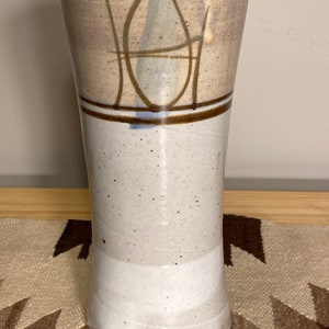 May include: A white ceramic vase with a brown and blue abstract design. The vase has a speckled glaze and a rounded bottom.