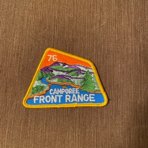 May include: A triangular shaped embroidered patch with a yellow border. The patch features a mountain scene with a river and the text "Campore Front Range" and the number "76".
