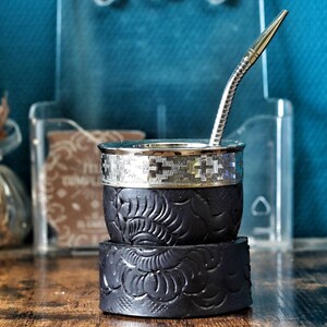 Peut inclure: Une calebasse à maté noire avec un motif floral texturé et un bord en métal argenté orné d'un design géométrique. Une longue paille bombilla en métal argenté est insérée dans le haut de la calebasse, qui repose sur une base assortie.