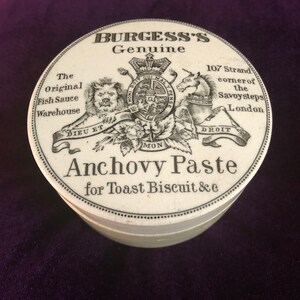 May include: A white ceramic jar lid with black lettering that reads "Burgess's Genuine" and "Anchovy Paste for Toast Biscuit &c". The lid also features a black and white illustration of a royal coat of arms with the text "107 Strand corner of the Savoy steps London".