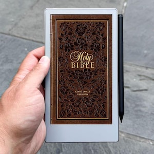 May include: An e-reader with a brown, ornate cover that reads "Holy Bible" in gold script. The e-reader has a black stylus and is held against a gray background.
