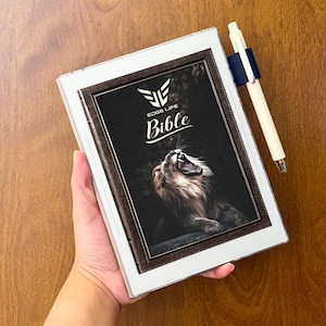 May include: A white digital e-reader device featuring a dark brown leather-textured cover with a graphic of a roaring lion. The screen displays the text "EDGE LIFE Bible" above the lion. A cream-colored pen is attached to the side with a blue clip.