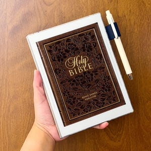 May include: A brown leather-bound Holy Bible King James Version with an intricate floral embossed cover, encased in a white protective tablet-style frame, accompanied by a cream-colored pen held in a dark blue elastic loop attached to the side.
