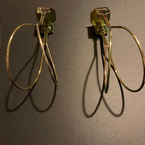 May include: Two gold metal curtain tiebacks with a simple, looped design. The tiebacks are attached to a wall with a screw.