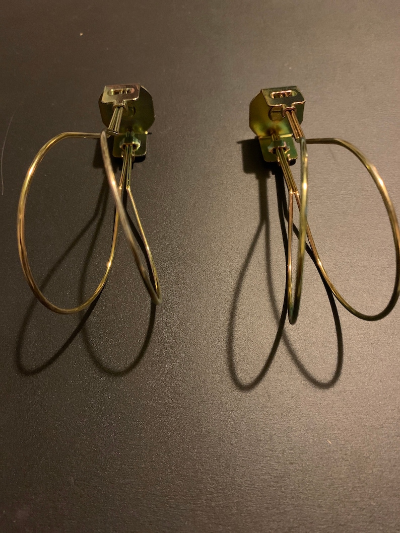 May include: Two gold metal curtain tiebacks with a simple, looped design. The tiebacks are attached to a wall with a screw.