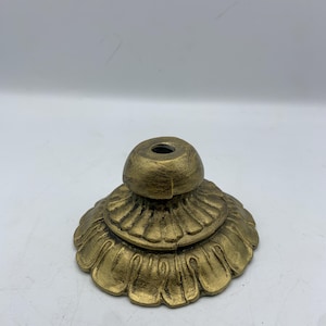 May include: A gold-colored, decorative base with a floral design. The base has a tiered structure with a rounded top and a central hole. The base is made of metal and has an aged, antique appearance.