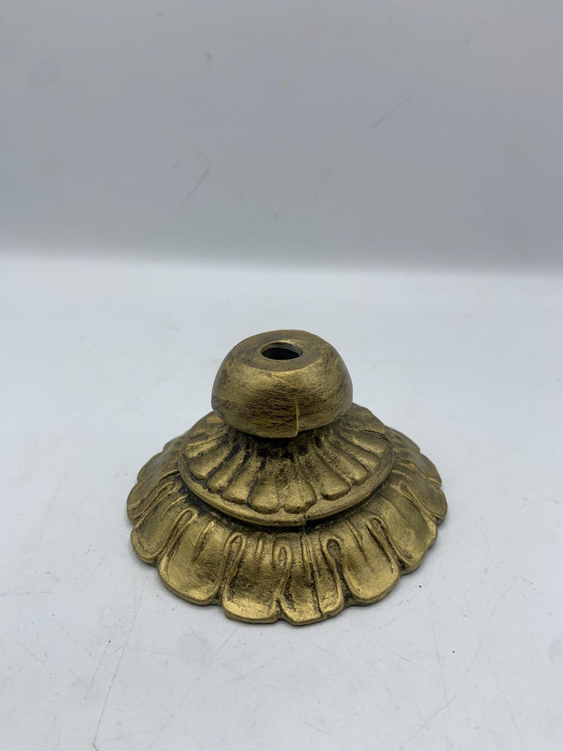 May include: A gold-colored, decorative base with a floral design. The base has a tiered structure with a rounded top and a central hole. The base is made of metal and has an aged, antique appearance.